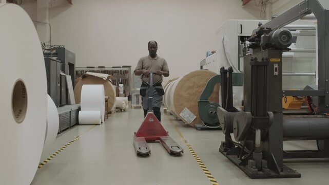 Long shot of mature Black man working in printing house walking along workshop pushing pallet jack