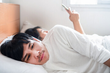 Asian couple having marriage problems at home, husband lying in bed while wife looks at his smartphone, showing family conflict, cheating, arguments, stress, and relationship issues leading to divorce