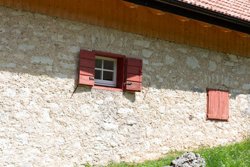 Sunlight casts strong shadows across a weathered stone wall and red window shutters, evoking a warm, peaceful summer afternoon in a rural village.