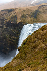 The majestic Skogafoss waterfall in Iceland cascades down a steep cliff surrounded by green meadows at the top of the waterfall against cloudy sky