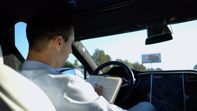 Male businessperson reading book during riding on electrical vehicle with autopilot at urban road. Successful businessman improving his knowledge while riding an autonomous self driving electric car - Powered by Adobe