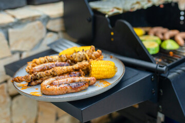 Grilled sausages, skewers, and corn on a plate near a gas barbecue with vegetables and flatbreads cooking. Outdoor summer cooking scene with delicious grilled food.