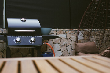 Close-up of a wooden outdoor table with a blurred gas grill in the background. Backyard BBQ setup ready for summer cooking and social gatherings.