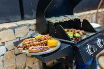 Grilled sausages, skewers, and corn on a plate near a gas barbecue with vegetables and flatbreads cooking. Outdoor summer cooking scene with delicious grilled food.