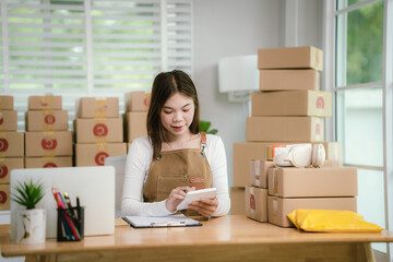 A young Asian entrepreneur happily works in her office, managing online retail orders and shipping packages, using technology to make her startup successful and grow.