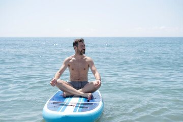 Fototapeta premium Man meditating on paddle board in ocean practicing yoga