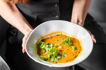 Close-up of hands holding a white bowl with creamy orange soup, garnished with cilantro and green onions, against a dark background.