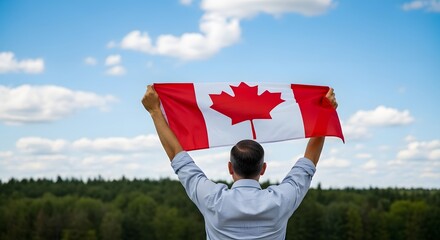 Man holding the canadian flag high in the air against a blue sky with clouds and a green forest background, representing patriotism and national pride