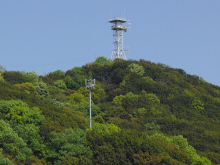 At the top of the mountain, an antenna tower and a large parabolic antenna can be seen. Lower down, a mobile phone antenna serves the residential area below.