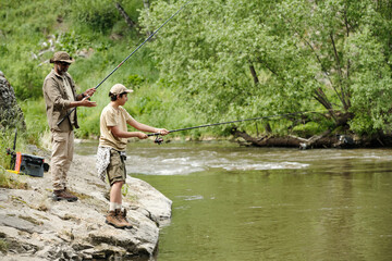 Middle aged man and boy fishing together on rocky riverbank, both holding fishing rods and focusing on water, lush green trees and flowing river in background