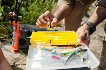 Middle aged man holding fishing lure and open tackle box with various fishing baits outdoors, preparing equipment for fishing activity with child or son nearby