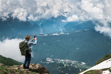 Young man traveler with backpack standing on the top of the mountain and taking photo with smartphone