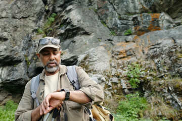 Middle aged South Asian man standing outdoors near rocky cliff checking smartwatch while holding fishing rod, wearing backpack and cap, preparing for fishing trip with son