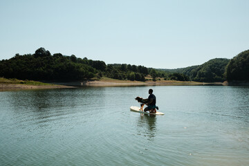 A man paddling on a SUP board with his Australian shepherd dog on a calm lake. Summer outdoor recreation and pet adventure