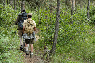 Fototapeta premium Middle aged man and boy hiking together through forest trail carrying backpacks and fishing rods, walking side by side surrounded by dense green trees