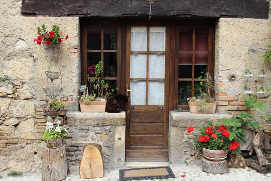 Rustic stone house entrance with wooden door, red flowers, and potted plants, featuring a black cat sitting by the window. A charming scene of countryside life and cozy home decor. Farfa, Lazio, Italy