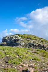 A scenic view of a rocky green hillside with layers of stone cliffs and fresh vegetation, set against a bright blue sky with scattered clouds. Perfect for themes of nature, geology, travel, adventure