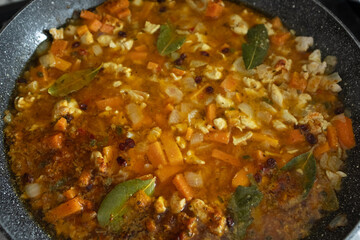 A close-up, high-angle shot of rice, spices, and other ingredients submerged in a rich broth during the cooking process. The vibrant orange liquid and visible white rice grains highlight a key step