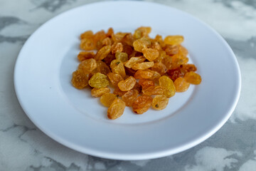 A pile of golden raisins sits on a small white plate against a marble background. These sweet, dried grapes are a traditional Plov ingredient, adding a unique flavor. Perfect for recipe blogs and cook