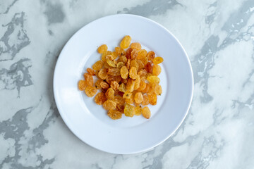 A pile of golden raisins sits on a small white plate against a marble background. These sweet, dried grapes are a traditional Plov ingredient, adding a unique flavor. Perfect for recipe blogs and cook