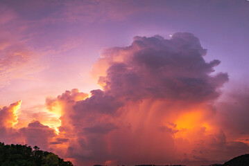 Fototapeta premium A vivid and dynamic sky captures towering storm clouds illuminated by the warm hues of a setting sun, creating a striking contrast between light and shadow above a darkened forested landscape.