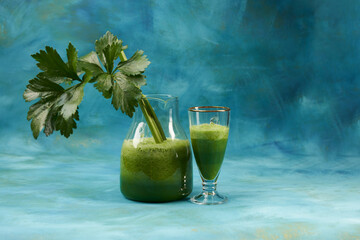 Glass pitcher and glass goblet with green thick smoothie on blue textured background. Celery stalk in the pitcher. Copy space.