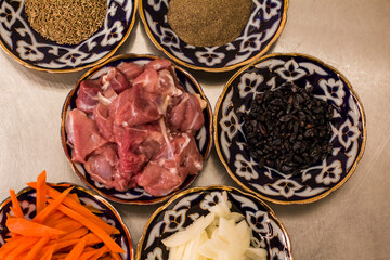 Colorful assortment of fresh ingredients for traditional Uzbek cuisine, pilaf, featuring spices, vegetables, and meat arranged in decorative bowls on a kitchen surface. Copy space, menu
