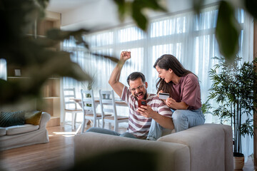 Happy couple celebrating online shopping success with credit card and smartphone at home