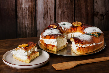 A slice of Roscon de Reyes filled with cream on a plate, with the rest of the Roscon in the background, on a wooden table.