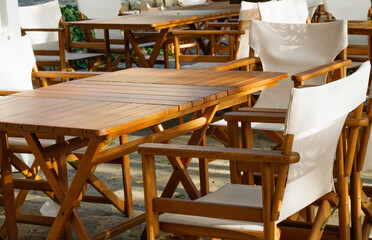 Wooden tables and chairs with white fabric seats in outdoor café terrace in warm sunlight