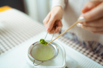 Asian woman preparing traditional Japanese matcha tea at home using bamboo tools, sifting matcha powder with care and precision.