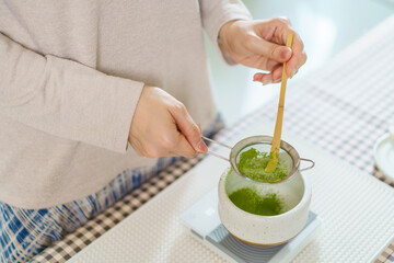 Asian woman preparing traditional Japanese matcha tea at home using bamboo tools, sifting matcha powder with care and precision.