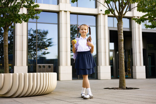 A young schoolgirl stands on the school yard, holding an apple, with a backpack, looking at the camera.
