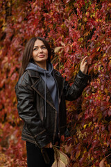 Beautiful young brunette girl walks in autumn park against the background of a wall with red leaves. autumn concept. autumn season. fall