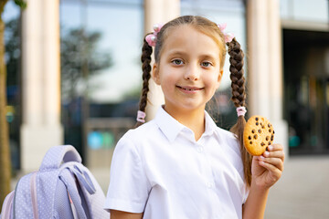 A Caucasian schoolgirl with braids smiles while holding a chocolate chip cookie on the schoolyard.
