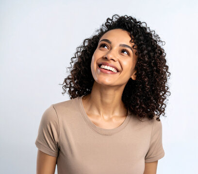 Mulher com cabelos cacheados, sorrindo e olhando para cima. Blusa bege. Fundo branco. 