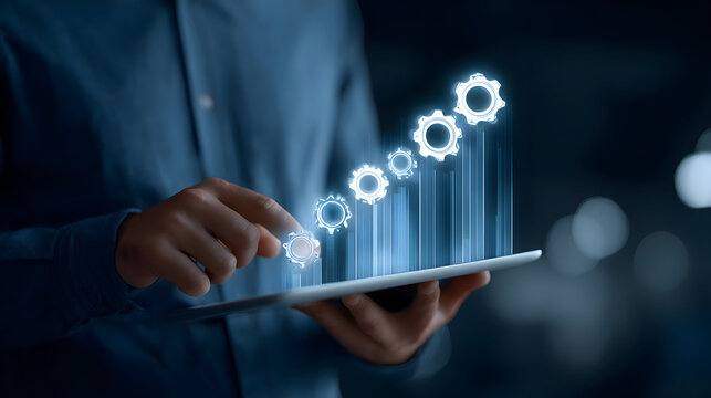 A business man holds a tablet with a glowing white gear icon, a transparent blue data graph rising around it.