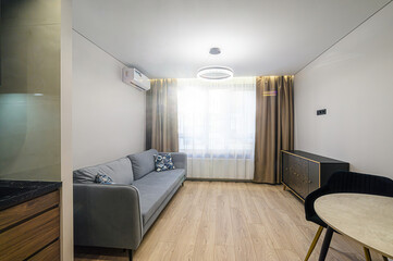 modern living room with a grey sofa, round table, sideboard, and light wood floors. The room is bright, lit by a ring light fixture and natural light through draped curtains