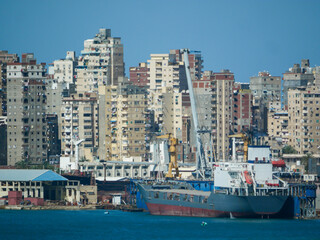 Alexandria, Egypt - 05.04.2025  Cargo ship docked at Alexandria shipyard with cranes, workshops, and high-rise apartment buildings in the background along the Mediterranean waterfront