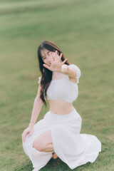 Fototapeta premium Embracing Sunlight: A young woman in light attire is posing in a lush green field, using her hand to shield herself from the sun, showcasing a moment of natural beauty and playful interaction.
