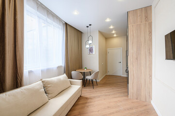 bright room showcases a beige sofa, a small dining table with two chairs, and wood flooring. A window with curtains provides natural light. Recessed lighting illuminates the scene