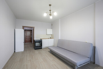 Fototapeta premium simple studio apartment. Features a sofa, kitchenette with stove, and a fridge. Walls are light gray, floor is laminate