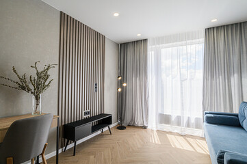 Modern room with wood slat wall, herringbone floor, grey curtains, blue sofa, and desk. Natural light streams in