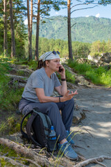An adult attractive woman is sitting on the rocks on the bank of a mountain river and talking on the phone.