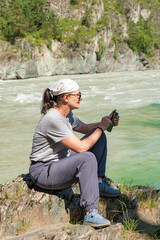 An adult attractive woman sits on the rocks on the bank of a mountain river and types a message on her smartphone