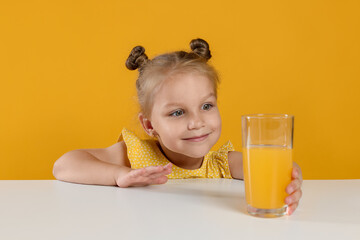 Cute little girl reaching for glass of juice at white table against orange background