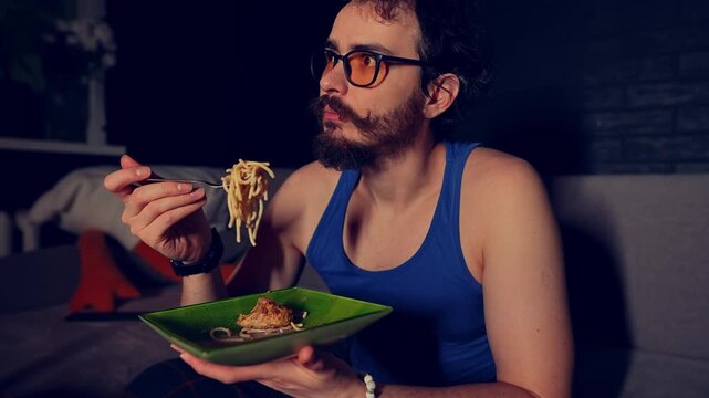 Closeup view of young adult guy sitting at home alone at night and eating pasta with chicken while watching a movie in living room. Slowmo of Caucasian man having tasty homemade dinner in front of TV.