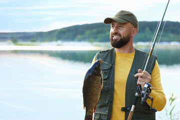 Smiling fisherman holding rod and caught fish near lake at summer. Space for text