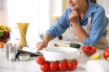 Senior woman using tablet while cooking at white marble table in kitchen, closeup