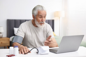Fototapeta premium Senior man measuring blood pressure while having online consultation with doctor via laptop at table indoors